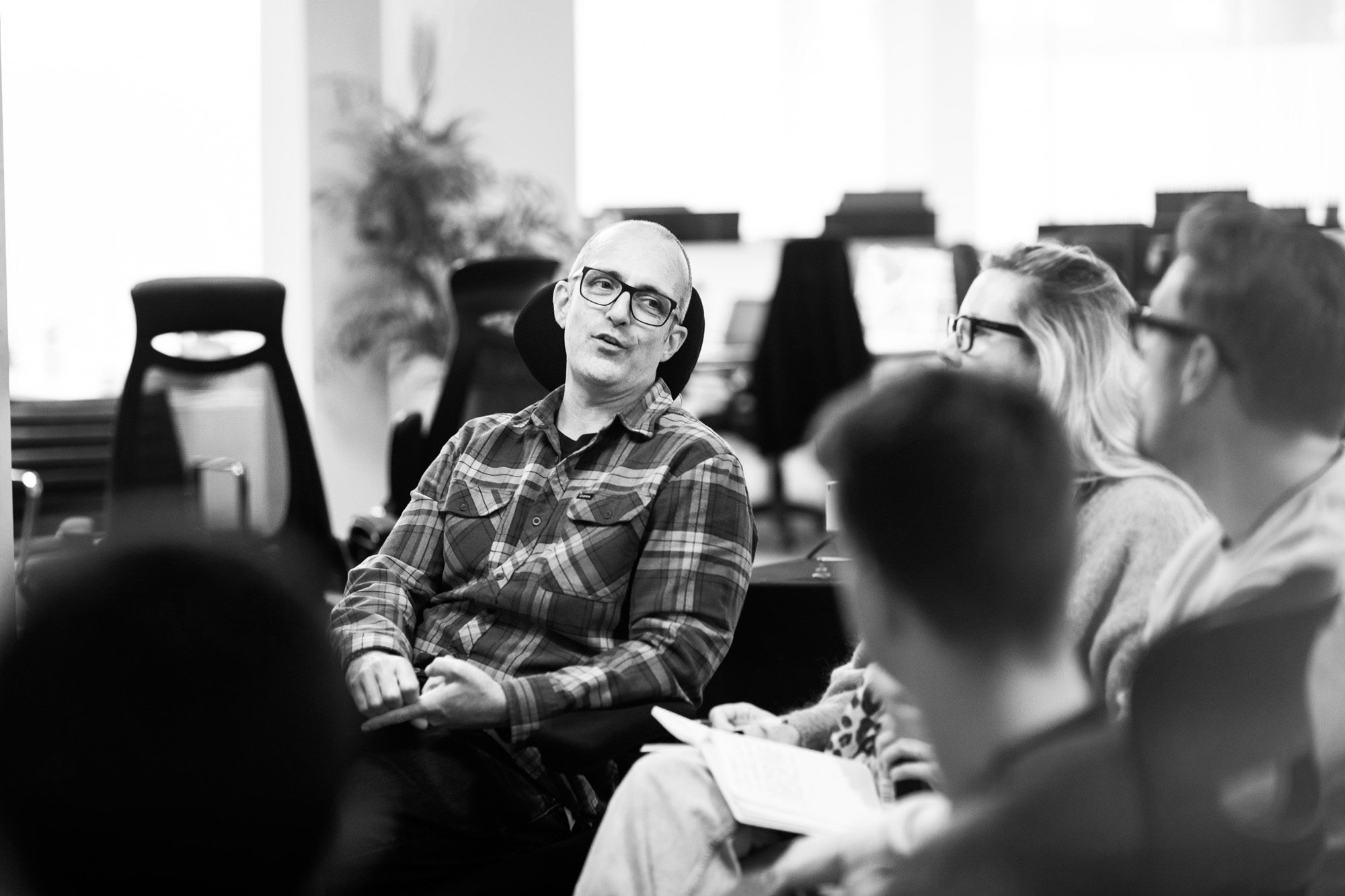 Bald man in plaid shirt and glasses sitting in office meeting with colleagues in modern workspace