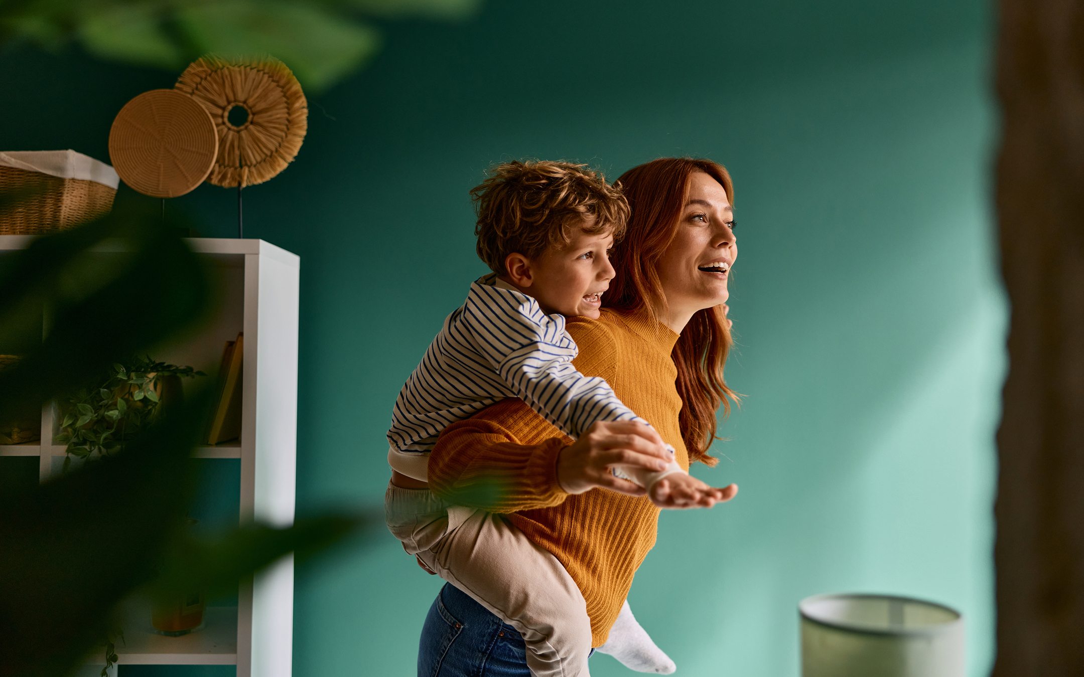 Mother giving piggyback ride to young son in modern teal living room, both smiling and looking forward