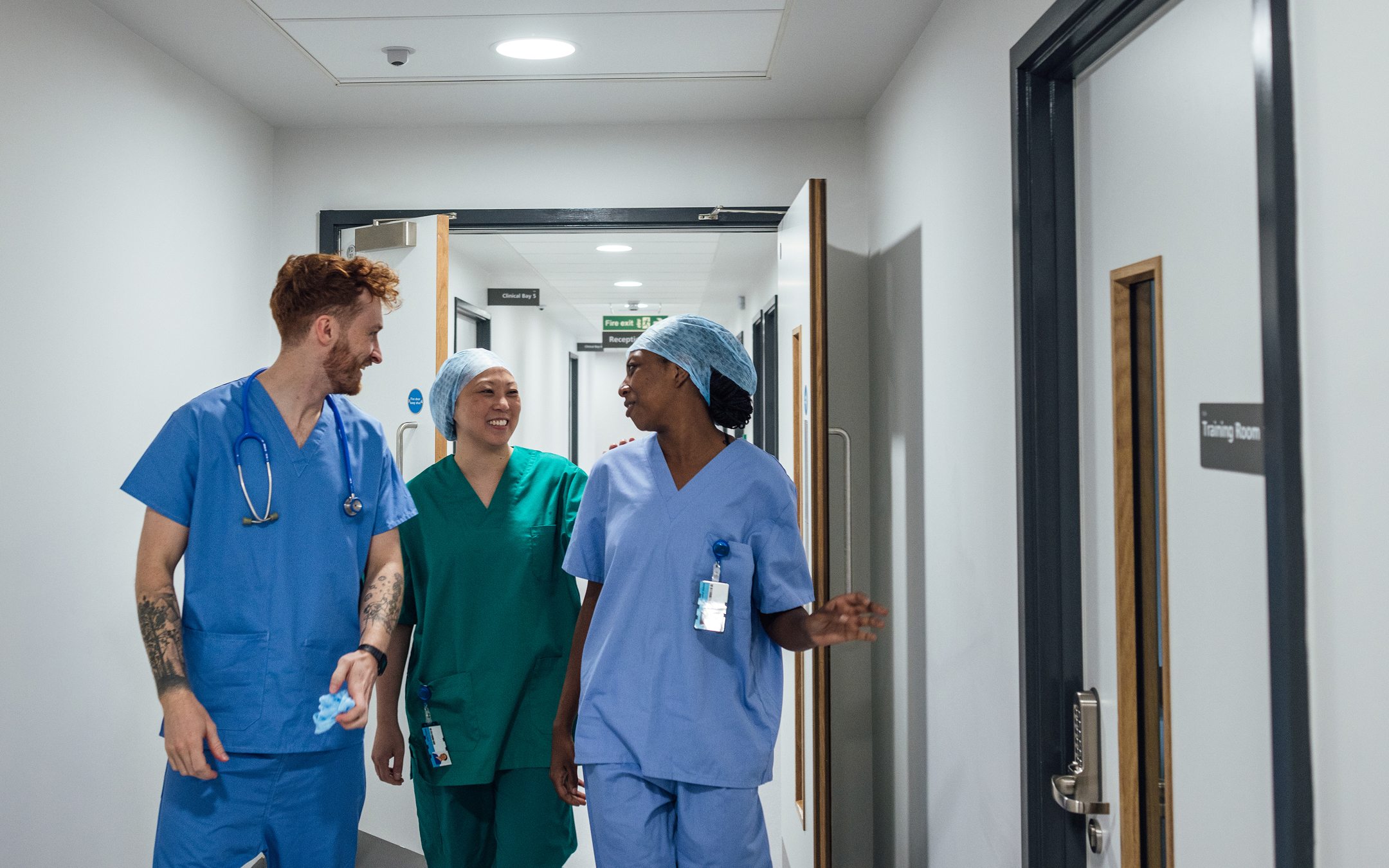 Three healthcare workers in scrubs walking together down a hospital corridor, smiling and conversing
