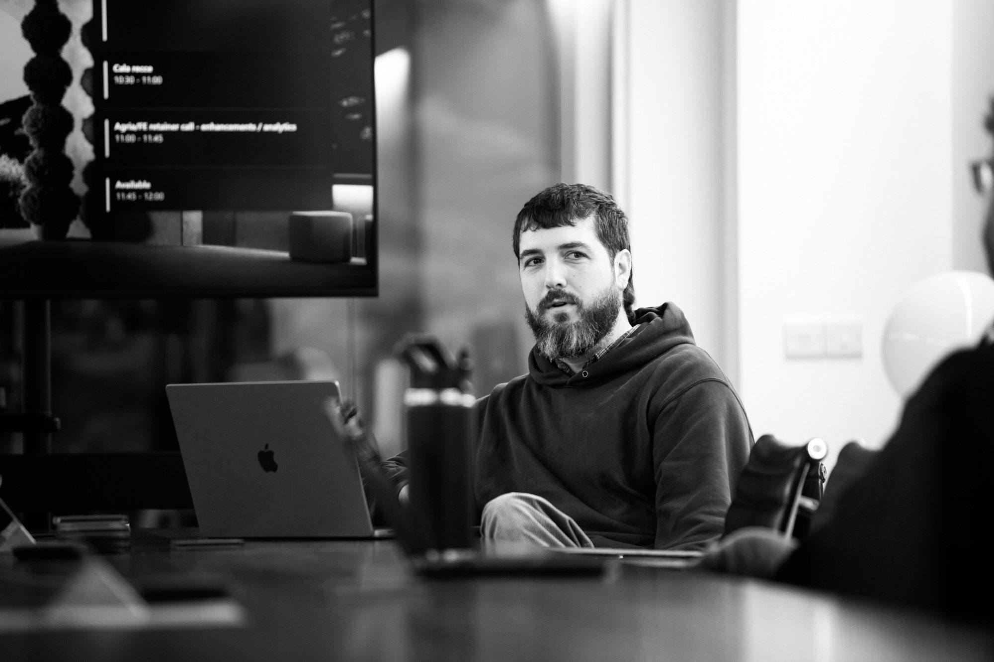 Bearded man in hoodie working on laptop at modern office desk in black and white