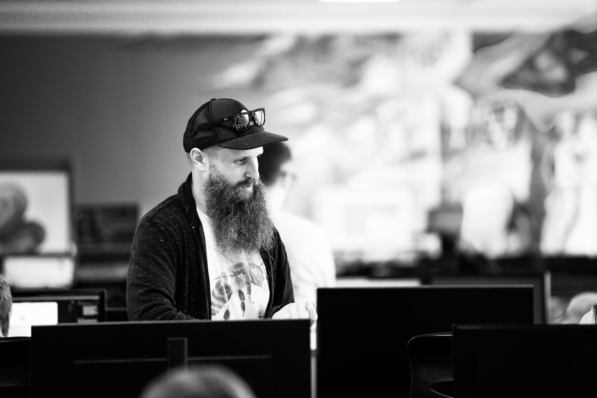 Bearded man in cap and hoodie working at computer in modern office environment