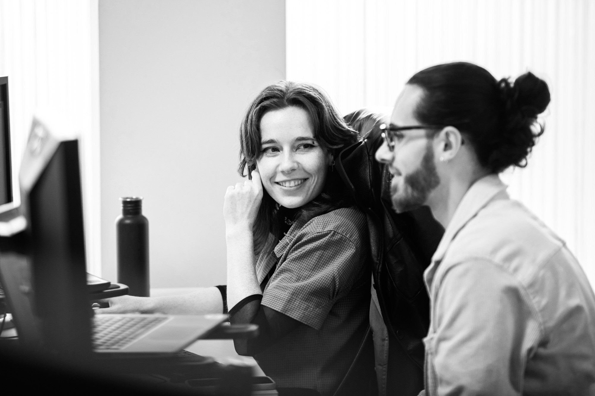 Two professionals smiling while working together at a desk with laptop in modern office setting
