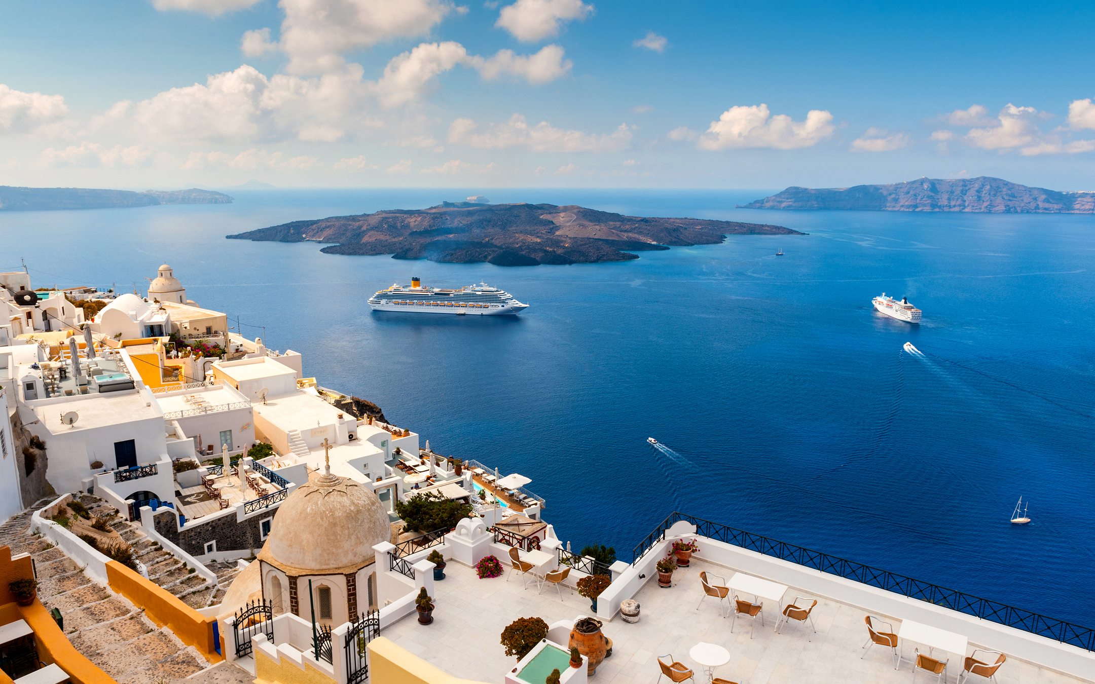 Santorini clifftop village with white buildings overlooking blue sea, cruise ship and volcanic island in distance