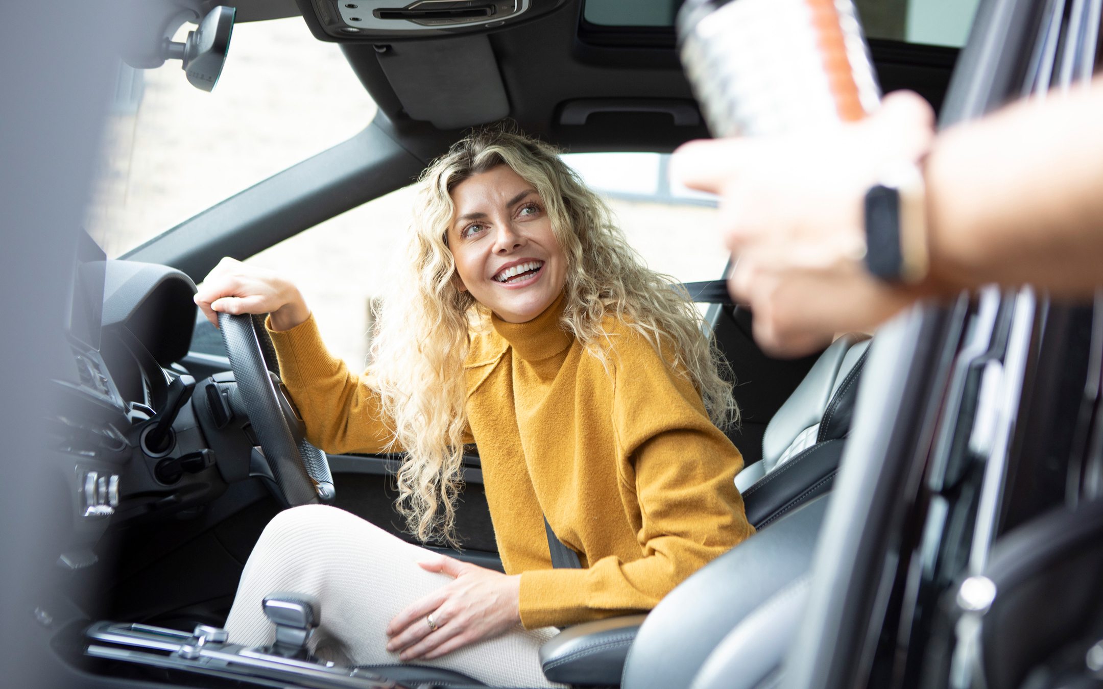 Happy blonde woman in yellow sweater sitting in car driver's seat receiving car keys from hand reaching into frame