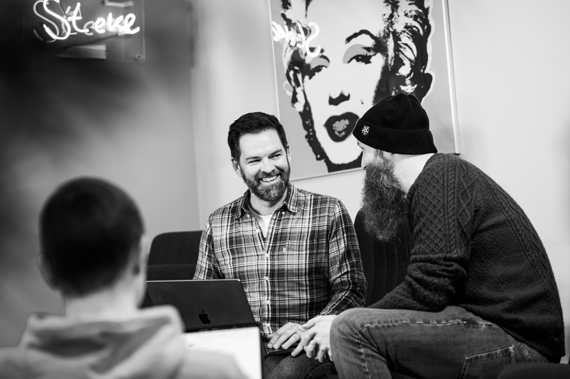 Two men collaborating at a laptop in a modern office, one wearing plaid shirt, one in beanie and sweater, black and white photo