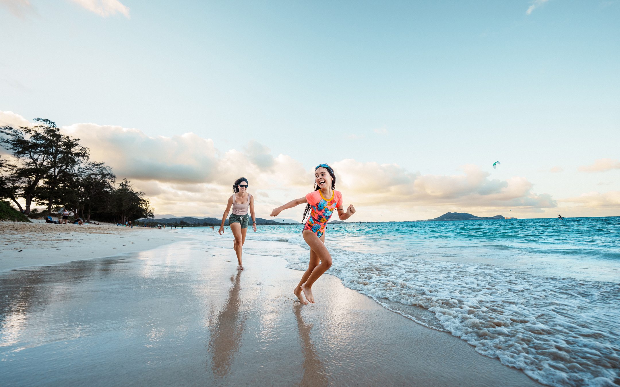 Two women running on tropical beach at sunset with turquoise water and mountains in background